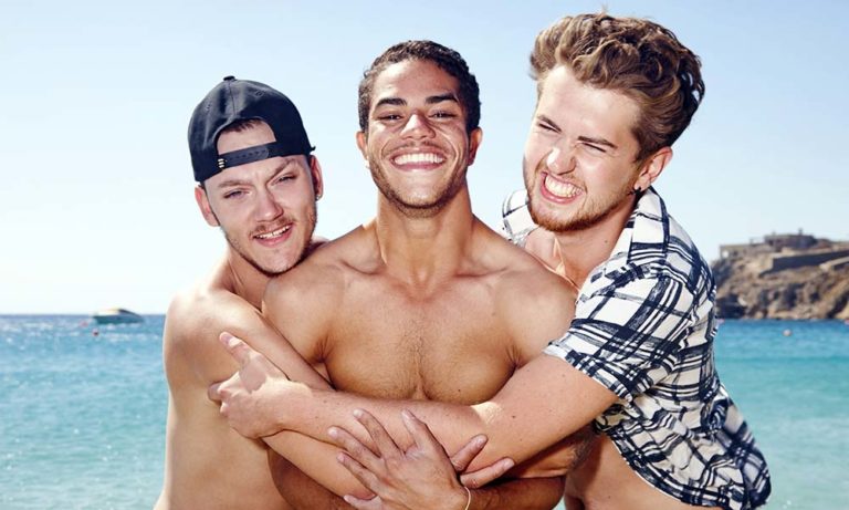Three shirtless men hug on a beach in Mykonos. Gay travel, summer vacation, and reality TV show concept. Blue sea and sky background.