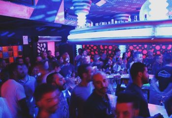 Inside Parthenon Gay Cruise Club in Torremolinos. Crowded bar scene with men under blue & pink lights. Gay nightlife travel.