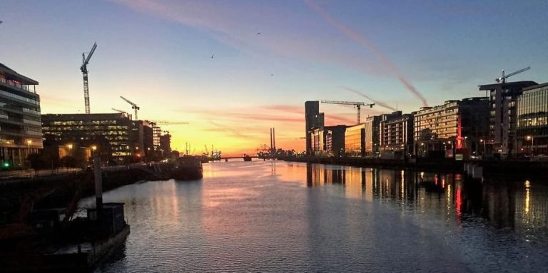 Sunset over Dublin's River Liffey, seen on a gay-friendly bike and e-bike tour. Cityscape with cranes, buildings, and water reflections.