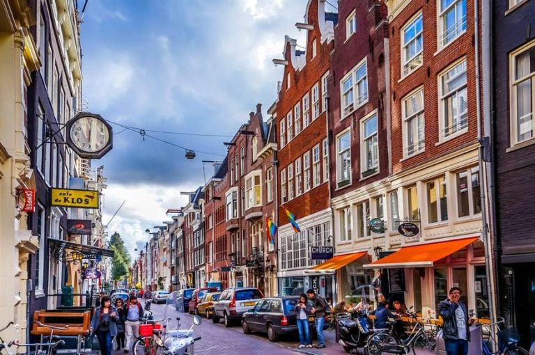 Amsterdam's Red Light District coffee shop tour: Canal street view with cafes, shops, cars, bikes, and people. Gay travel advice.