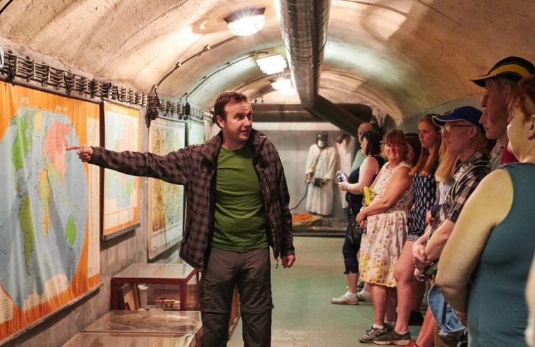 Prague's Communist history: A tour guide points to a map in a nuclear bunker. Tourists listen intently. Cold War history exploration.
