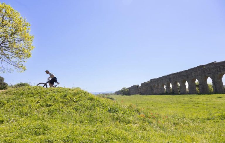 Gay man's Appian Way E-Bike Adventure: Cyclist on grassy hill near Roman aqueducts. Rome travel, catacombs, and Roman bites.