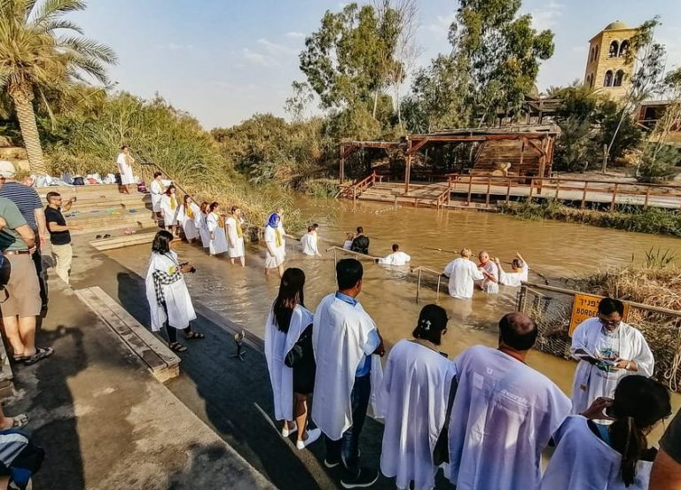 People baptizing in Jordan River during a day trip to sacred sites like Bethlehem & Jericho. Scenic view with greenery & architecture.