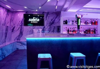 Inside Sauna Sitges, a gay sauna in Sitges, Spain. Bar area with stools, illuminated bar, and shelves of liquor bottles. Purple lighting.