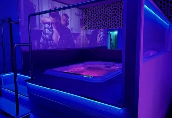 Interior of a gay sauna in Sitges. A hot tub glows with pink light, surrounded by blue LED strip lighting. A screen shows two men embracing.