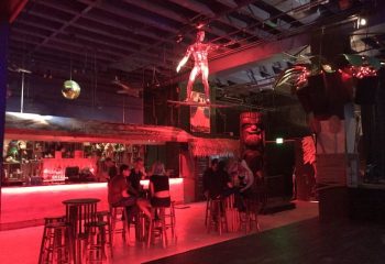 Scarlet Honolulu gay bar interior. Patrons seated at high-top tables, tiki decor, and a surfer statue overhead. Red lighting.