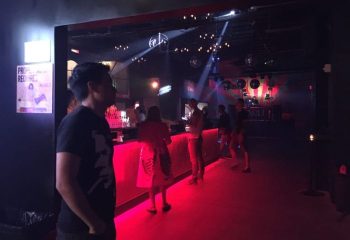 Inside Scarlet Honolulu gay bar, patrons stand near the red-lit bar. Disco balls hang above. Honolulu gay nightlife scene.