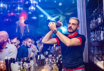 Bartender at Shamone Club, a gay bar, mixes drinks. Disco balls, blue lighting, and a crowd add to the vibrant nightlife atmosphere.