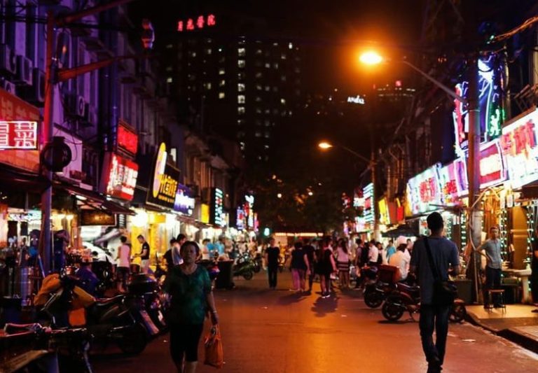 Gay-friendly Shanghai street food tour. Busy street at night with neon signs and people. Dumplings and desserts are popular bites.