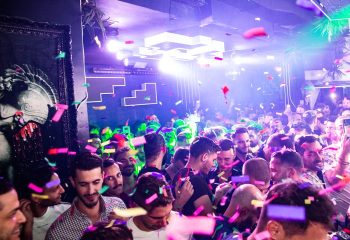 Sodade2: Gay men celebrate at a crowded, vibrant party with confetti and bright lights. Nightlife travel scene for gay men advice.