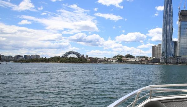 Sydney Harbour cruise view featuring the Harbour Bridge, city skyline, and modern architecture under a cloudy blue sky. Gay travel destination.