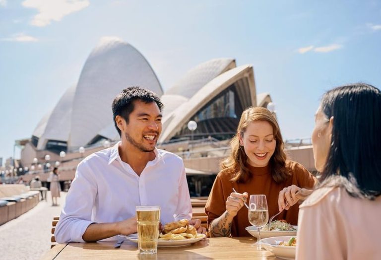 Sydney Opera House tour: People dining with harbor views. Culture, cocktails, and harborside dining experience. Gay travel advice.