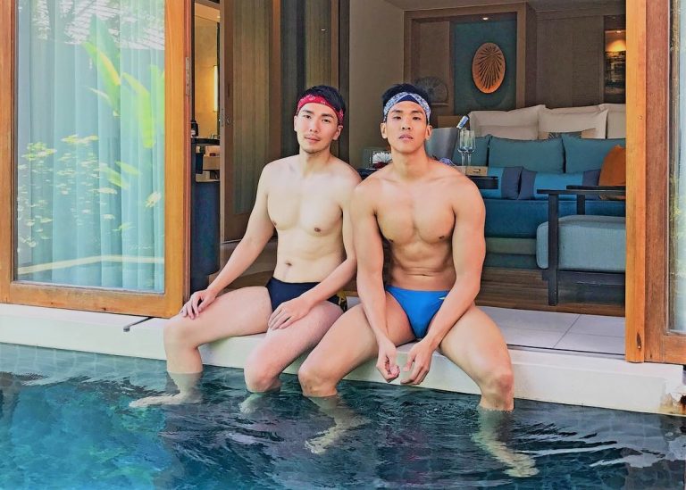 Gay couple in Phuket, Thailand, at Renaissance Phuket resort. Two fit men in swimsuits sit by a pool, enjoying their gay travel adventure.