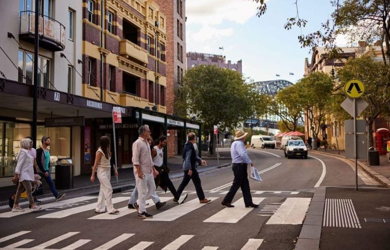 Sydney's The Rocks: People on a historic stroll at a crosswalk with iconic views of the Harbour Bridge in the background. Gay travel advice.