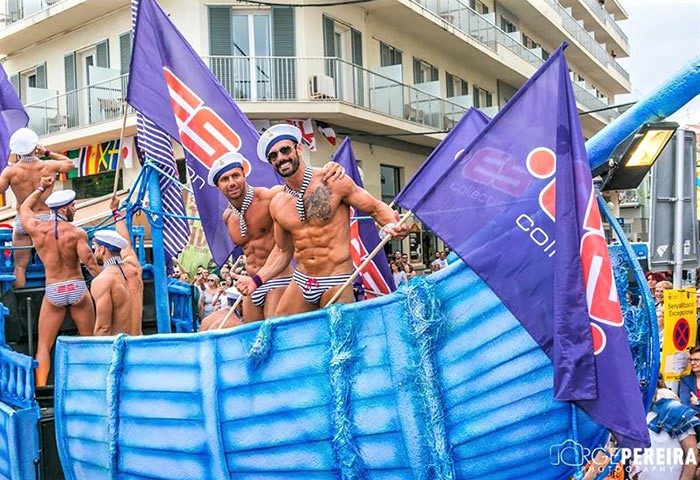 Gay Pride Sitges party: Shirtless men in sailor hats and striped swimsuits on a blue boat float, waving flags. Gay Sitges travel guide.