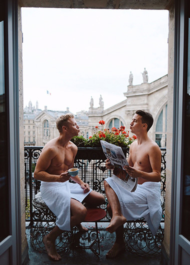 Gay honeymoon in Paris. Two shirtless men in towels on a balcony, one with coffee, one with a newspaper. Paris buildings in background.