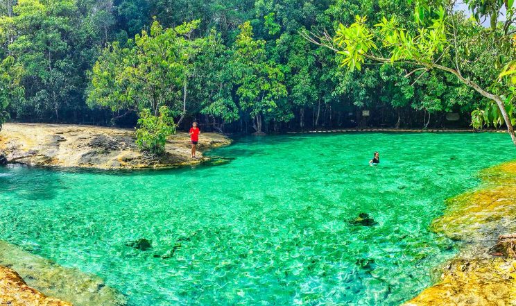 Thailand's Emerald Pool, perfect for gay adventure. Man stands on shore; another swims in the crystal-clear, hot spring water. Lush jungle backdrop.