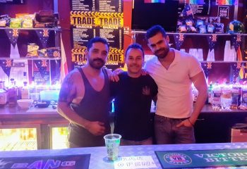 Three gay men pose at TRADE Club Sydney bar. Snacks, drinks, Australian flags, and Bar Open sign visible. Gay travel advice.