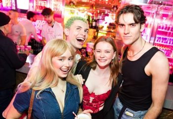 Sydney nightlife: Smiling group at Universal Sydney bar. Gay travel advice. Men, women, drinks, and vibrant pink lighting. Fun night out.
