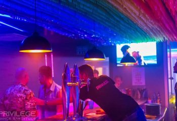 Vertigo Dance Bar, a gay club in Sitges. Bartender with Privilege shirt serves drinks under rainbow decor and neon lights. Gay travel.