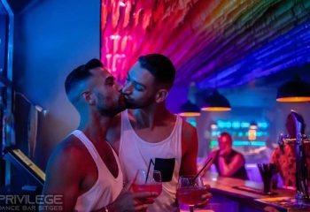 Gay couple kissing at Vertigo Dance Bar, a gay club in Sitges, Spain. Rainbow lights illuminate the dance floor. Gay travel destination.