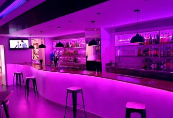 Interior of Vertigo Dance Bar, a gay club in Sitges, Spain. The bar is illuminated with purple lighting, stools line the bar.