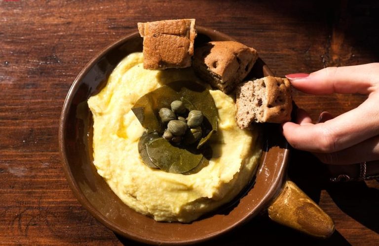 Local food in Santorini: Fava dip with capers, served with bread. A hand is holding a piece of bread. Best views and drinks on walking tour.
