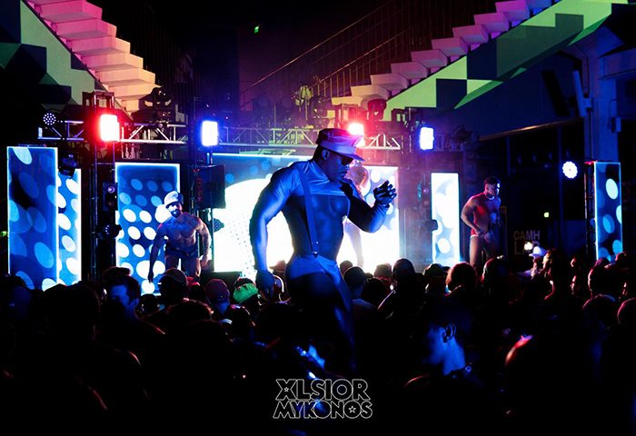 XLSIOR Mykonos party scene. Muscular men dance on stage at the gay event. Crowd enjoys the show with bright lights. Gay travel.