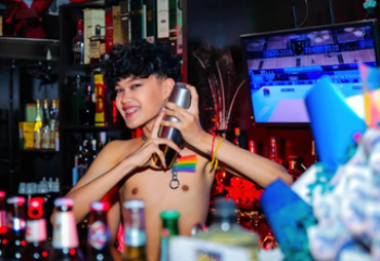Shirtless bartender at POC Gay Bar in Phnom Penh, Cambodia. Rainbow flag necklace, shaker, and bottles. Gay travel.