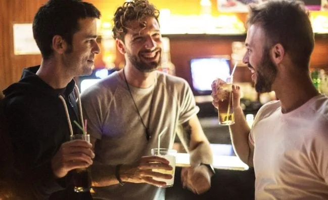 Three gay men laughing and holding drinks at a bar during a Madrid gay nightlife tour. Gay bar scene, travel advice for gay men.